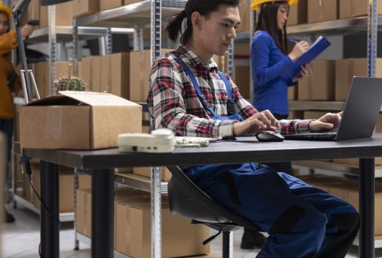 Asian staff member in a local brand supply room prepares products and taped boxes for shipment. Small business activity uses in house logistics for storage and delivery services.